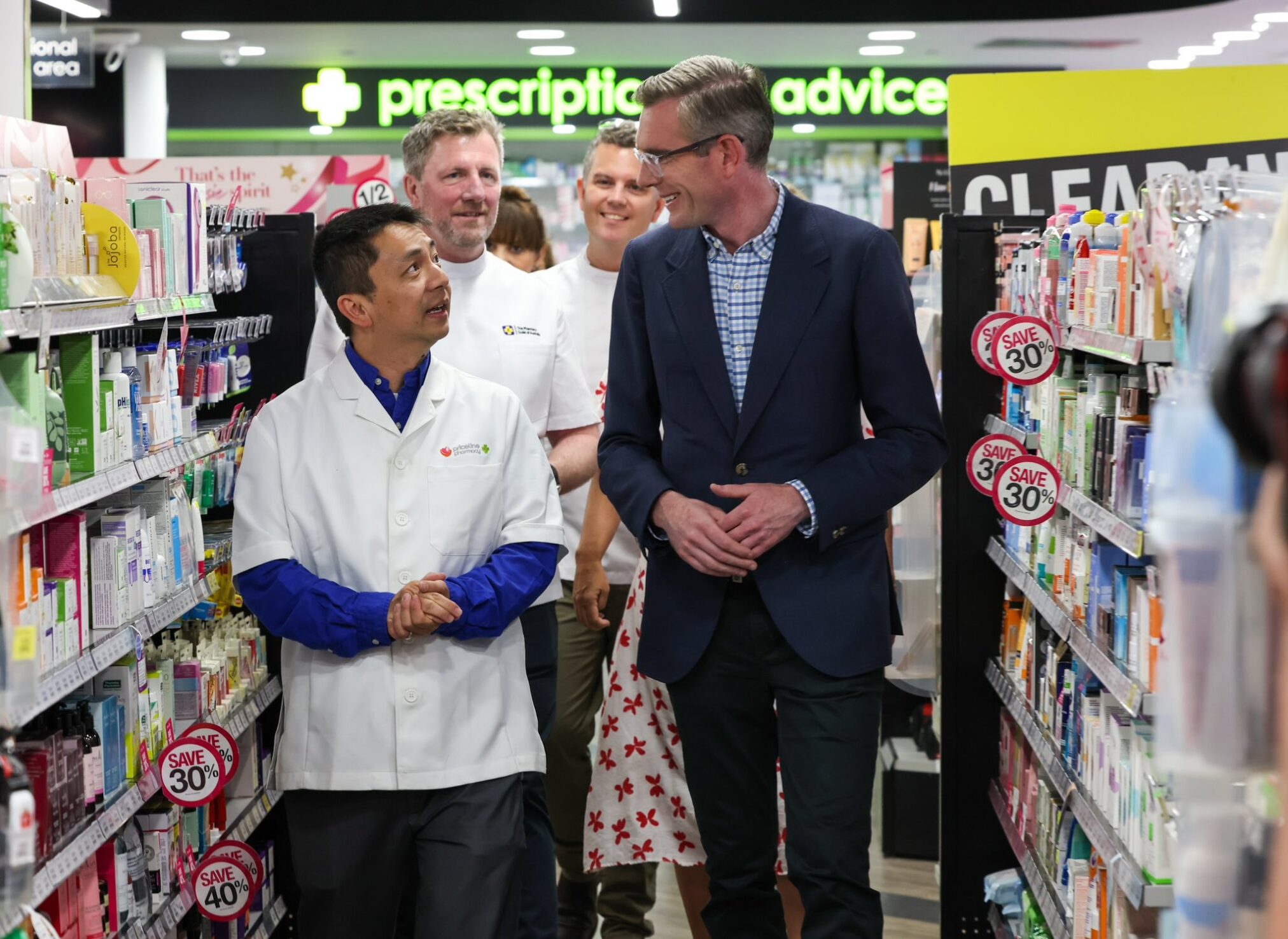NSW premier Dominic Perrottet in a pharmacy surrounded by shelves, talking to pharmacists